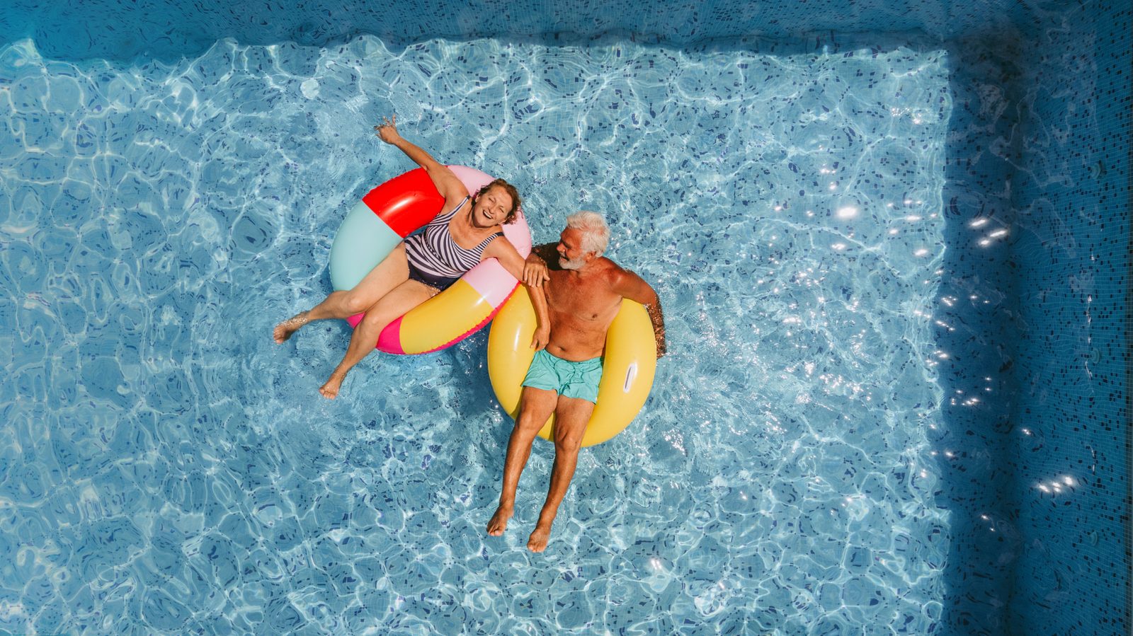 Older couple relaxing on inflatable pool floats in a swimming pool.