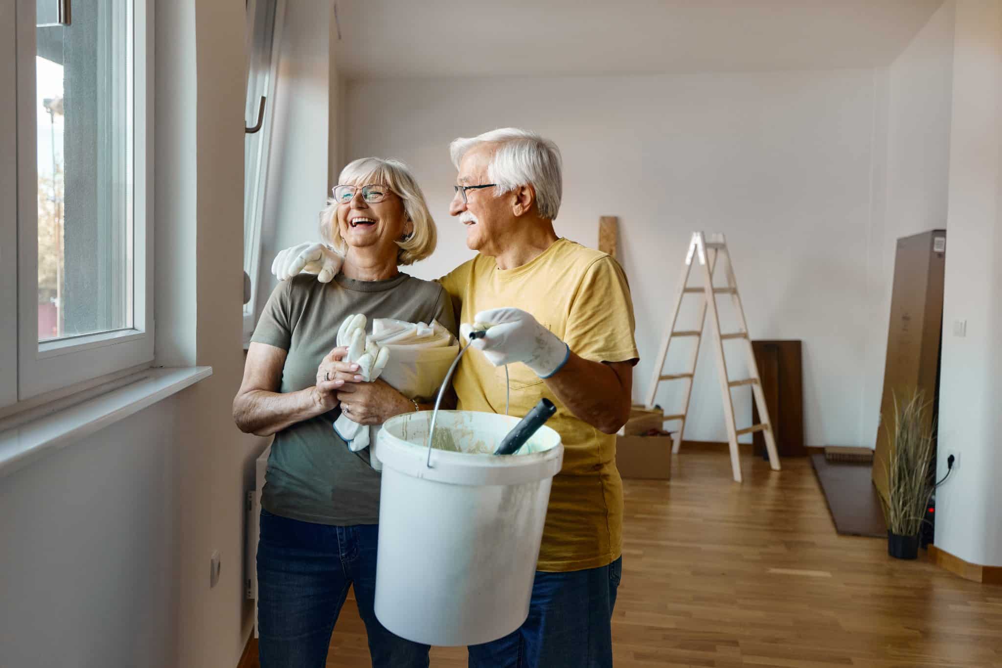 Couple holding a paint bucket and supplies in a room, smiling near a window.