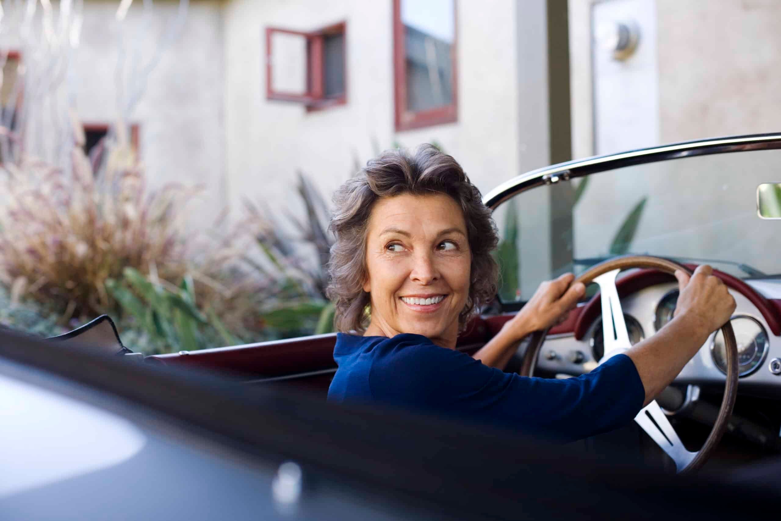 Older woman smiling while sitting in the driver’s seat of a convertible outside her home