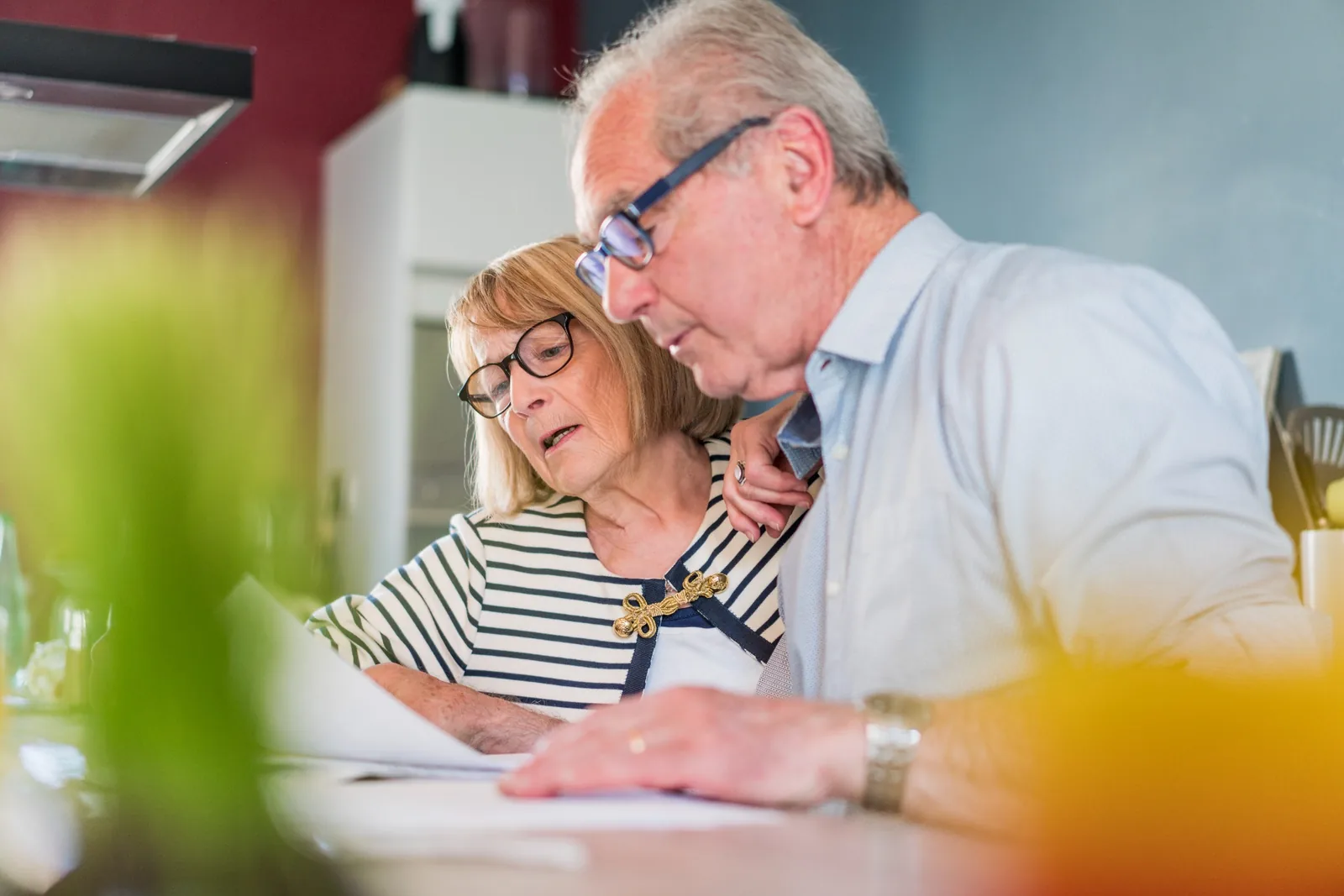 Older couple wearing glasses reviewing paperwork together at their kitchen table.